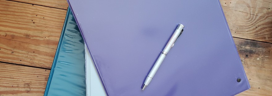 A pen rests atop a stack of binders on a rough wooden tabletop.