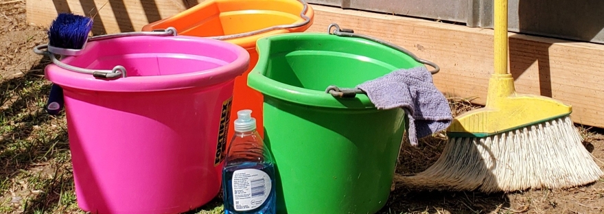 An assortment of cleaning supplies are arranged on the ground in front of a metal barn wall. The supplies include colorful buckets, a scrub brush, dish soap, and a broom.