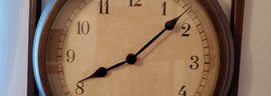 A clock with a decorative wooden frame hangs on a wall. It reads 8:07. Natural light shines on it from the left, but the room also seems to be lit with soft artificial light.