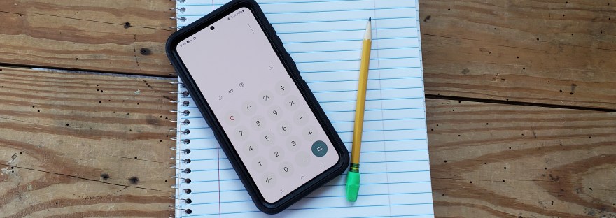 An open spiral notebook lies on a table. On top of the notebook are a pencil and a phone open to a calculator app.