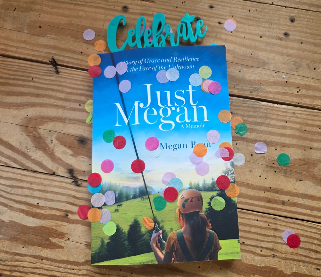 A copy of Megan Beanโs memoir, โJust Meganโ, rests on a rustic wooden tabletop, sprinkled with multicolored confetti. Painted wooden letters above it spell โCelebrateโ.